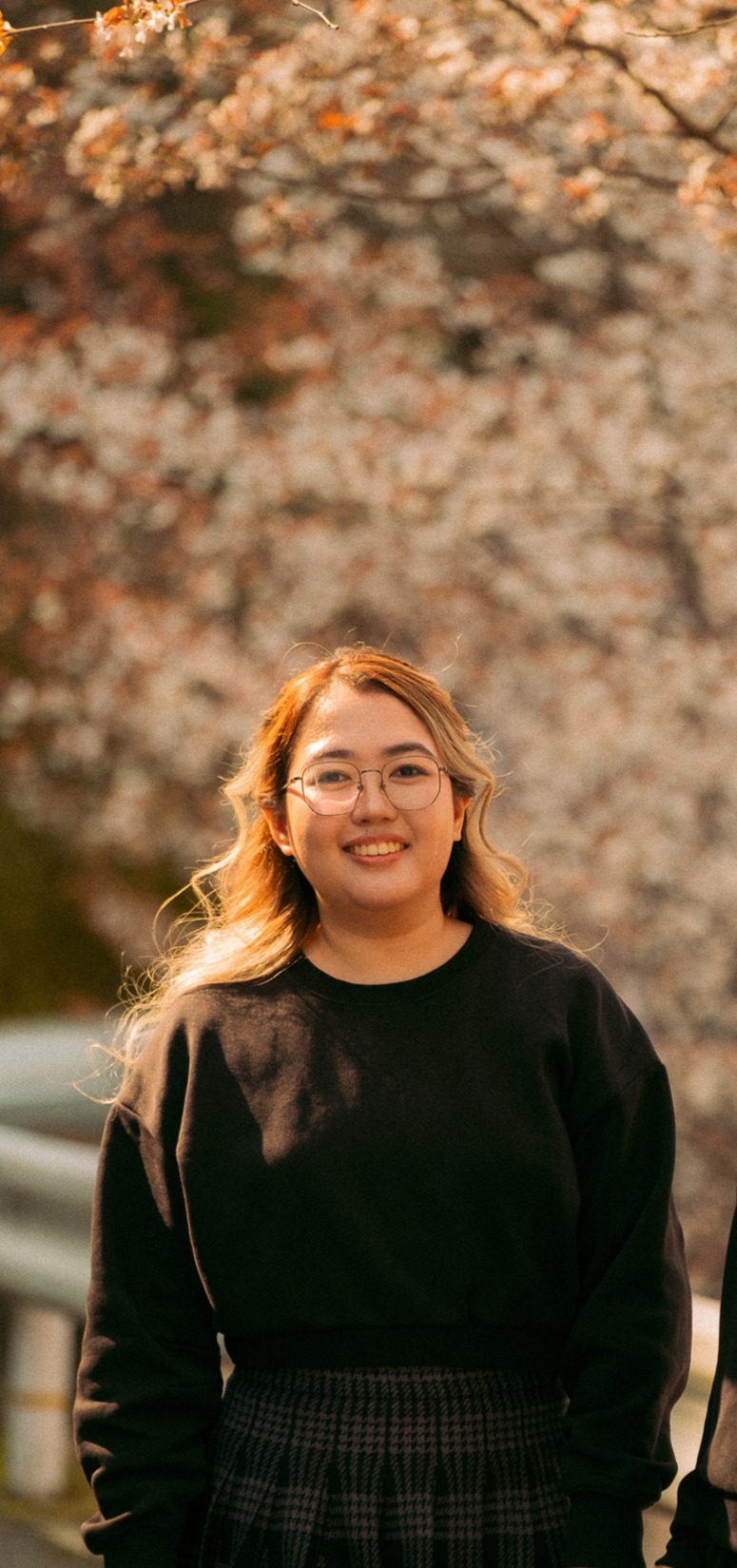 Mariane, smiling in a black sweater and glasses, standing under cherry blossoms in golden light.